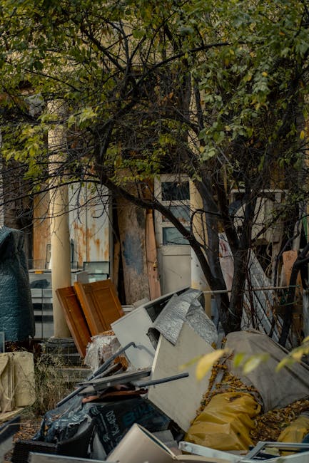 A cluttered outdoor area with a large pile of bulky renovation waste, including discarded wooden doors, electrical appliances, and construction debris, situated beneath a tree with dense green foliage. The surface of the waste items appears weathered, with some showing rust, peeling paint, and dust accumulation, indicative of recent demolition activity. The ground is covered with scattered materials such as plastic bags, cardboard, and other construction packaging. In the background, a building with a worn, rusted metal facade and partially visible windows can be seen. This scene emphasizes the importance of professional waste removal and deep cleaning services provided by Deep Cleaning Putney, highlighting their capability to manage bulky debris and maintain cleanliness after renovation projects.