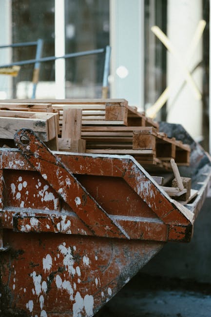 Close-up of an orange construction skip with white paint splatters, filled with wooden pallets and debris, positioned outdoors near a building with large glass windows. The pallets are weathered and stacked haphazardly inside the skip. Sunlight illuminates the scene, highlighting the textures of the wood and metal. This image relates to bulky renovation waste in Putney and showcases waste removal solutions offered by Deep Cleaning Putney, a professional cleaning service specializing in waste clearance for domestic and commercial properties, ensuring surfaces are free from debris and ready for subsequent cleaning or renovation work.