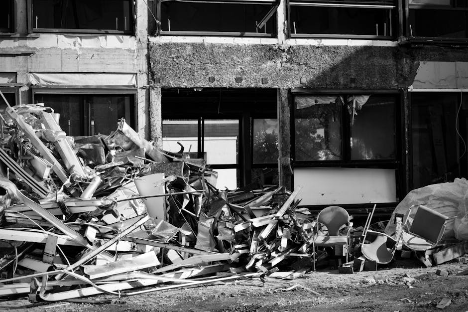 A cluttered outdoor area with a large pile of bulky renovation waste, including discarded wooden doors, electrical appliances, and construction debris, situated beneath a tree with dense green foliage. The surface of the waste items appears weathered, with some showing rust, peeling paint, and dust accumulation, indicative of recent demolition activity. The ground is covered with scattered materials such as plastic bags, cardboard, and other construction packaging. In the background, a building with a worn, rusted metal facade and partially visible windows can be seen. This scene emphasizes the importance of professional waste removal and deep cleaning services provided by Deep Cleaning Putney, highlighting their capability to manage bulky debris and maintain cleanliness after renovation projects.