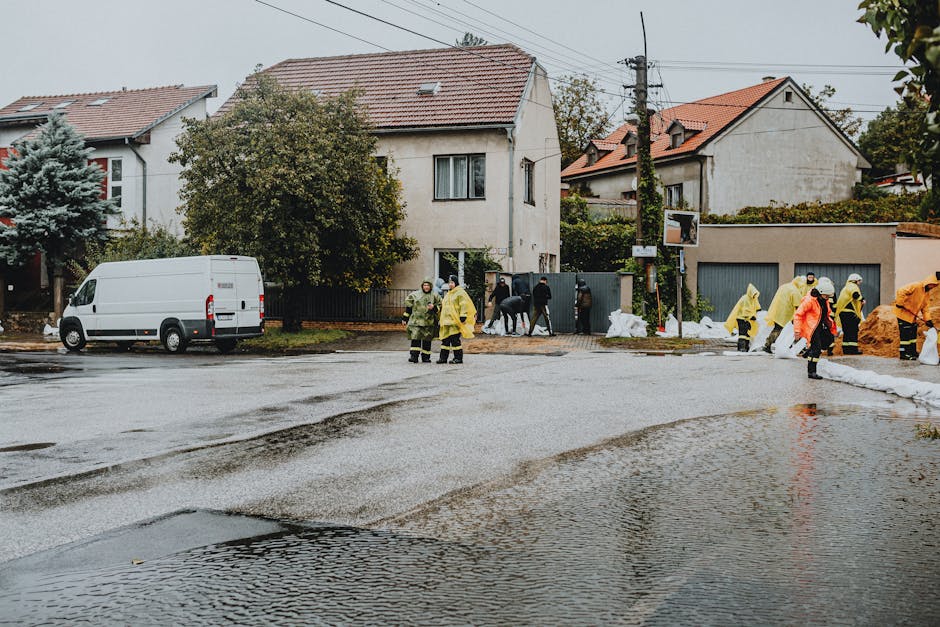 A residential street scene in Putney featuring multiple workers in yellow and orange protective suits managing flood cleanup. The street is partially flooded with water pooling on the cobblestone and asphalt surfaces. Several individuals are seen near a white van parked on the side, engaged in various cleanup activities. The background includes two-story houses with tiled roofs and trees, with some construction or cleanup materials visible. The scene is outdoors under overcast skies, with the area appearing wet and recently affected by flooding, highlighting the importance of thorough surface cleaning and sanitisation by specialists like Deep Cleaning Putney.