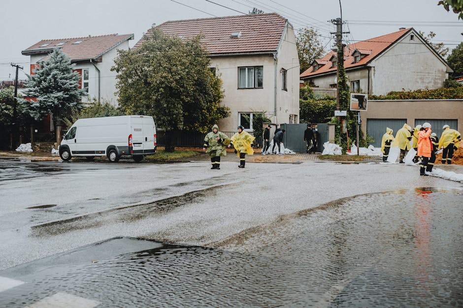 A residential street scene in Putney featuring multiple workers in yellow and orange protective suits managing flood cleanup. The street is partially flooded with water pooling on the cobblestone and asphalt surfaces. Several individuals are seen near a white van parked on the side, engaged in various cleanup activities. The background includes two-story houses with tiled roofs and trees, with some construction or cleanup materials visible. The scene is outdoors under overcast skies, with the area appearing wet and recently affected by flooding, highlighting the importance of thorough surface cleaning and sanitisation by specialists like Deep Cleaning Putney.