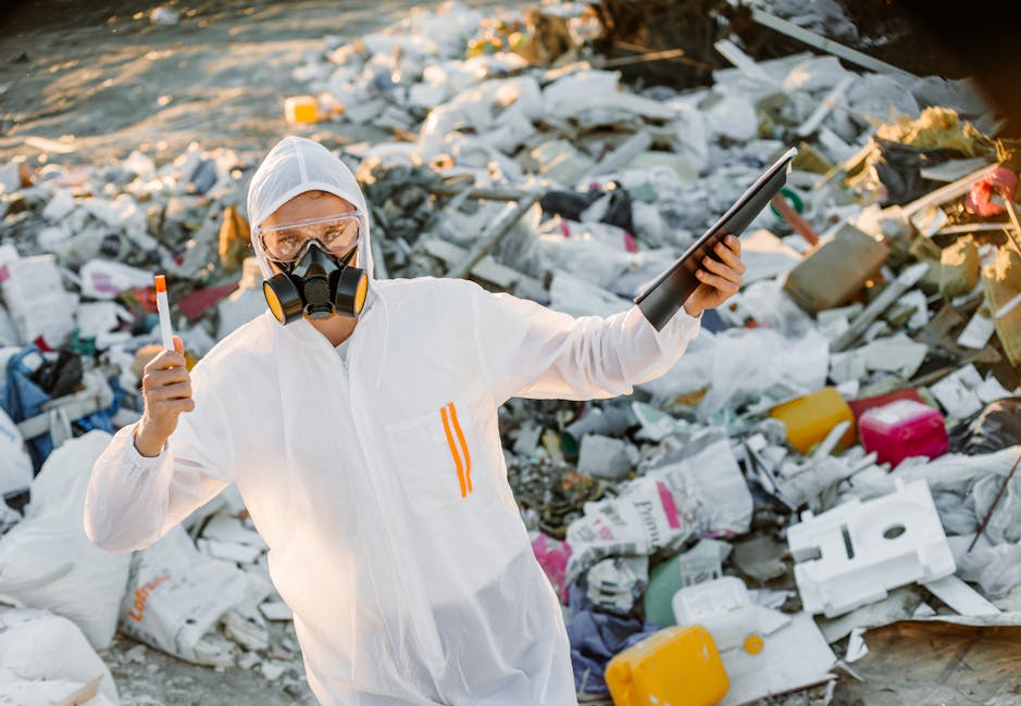 A person wearing a white protective suit, gloves, and a respirator mask is standing among a large pile of mixed household and industrial waste, including plastic bags, cardboard, and insulation materi
