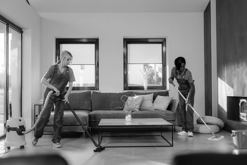 Two professional cleaning staff members from Deep Cleaning Putney stand back-to-back against a plain white wall, dressed in beige cleaning uniforms. One holds a vacuum cleaner, while the other is hold