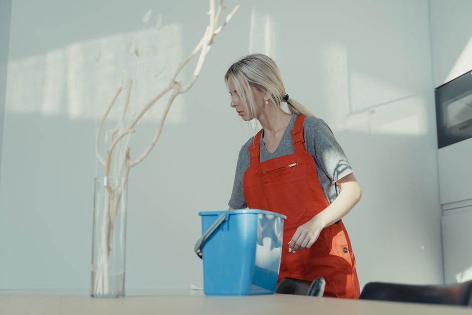 A woman with blonde hair tied back, wearing a gray t-shirt and a red cleaning apron, is applying surface cleaning and sanitisation in a bright, modern kitchen. She is standing next to a blue bucket, and appears to be wiping or preparing to clean a flat surface with cleaning tools or cloths. The kitchen features glossy gray cabinets and a stainless steel appliance, with natural light illuminating the scene, highlighting the cleanliness and shine of the surfaces. Deep Cleaning Putney offers reliable domestic cleaning and maintenance services, ensuring hygienic and well-maintained environments.