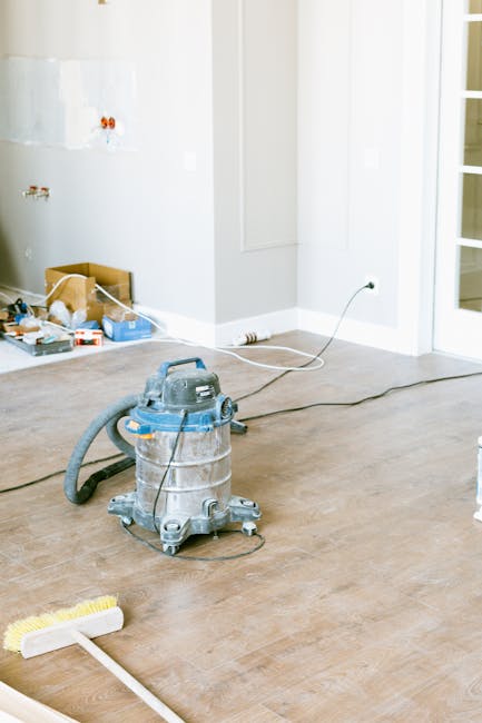 A woman with blonde hair tied back, wearing a gray t-shirt and a red cleaning apron, is applying surface cleaning and sanitisation in a bright, modern kitchen. She is standing next to a blue bucket, and appears to be wiping or preparing to clean a flat surface with cleaning tools or cloths. The kitchen features glossy gray cabinets and a stainless steel appliance, with natural light illuminating the scene, highlighting the cleanliness and shine of the surfaces. Deep Cleaning Putney offers reliable domestic cleaning and maintenance services, ensuring hygienic and well-maintained environments.