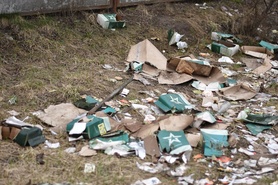 Waste and discarded cardboard boxes scattered across a grassy area, with some open and torn, alongside broken paper and packaging debris. The scene includes green cardboard boxes with star logos, partially flattened, surrounded by crumpled papers and torn cardboard pieces. The ground appears uneven with patches of grass and dirt, and there is no nearby structure visible. The overall impression is of unmanaged garden waste or litter, highlighting the importance of debris removal and proper waste disposal, which Deep Cleaning Putney offers through professional cleaning services. This image underscores the need for sanitary and well-maintained outdoor spaces in accordance with Wandsworth Council rules on garden waste and cleaning.